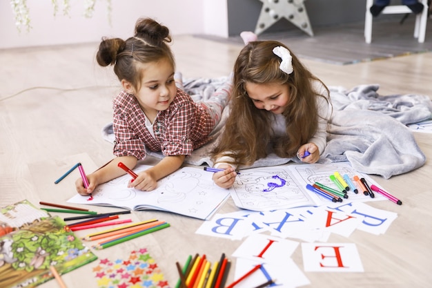Dos pequeñas muchachas felices que dibujan en el libro para colorear tirado en el suelo sobre la manta y aprendiendo letras