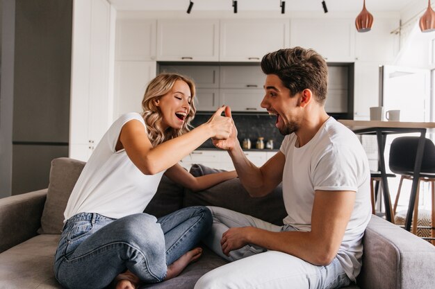 Dos personas alegres sentadas en el sofá y jugando con los pulgares. El hombre parece sorprendido y su novio se ve muy feliz y listo para ganar.