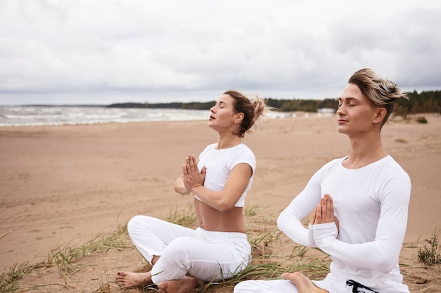 Dos personas europeas athelticas, hombre y mujer en ropa deportiva blanca cerrando los ojos y tomados de la mano en gesto namste, sentados en padmasana mientras meditan al aire libre durante un retiro de yoga junto al océano