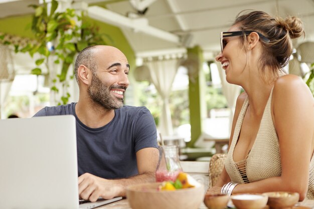 Dos personas felices divirtiéndose y riendo, sentados en la cafetería al aire libre durante el desayuno. Hombre alegre guapo con rastrojo con ordenador portátil, sonriendo y hablando con una mujer elegante en tonos.