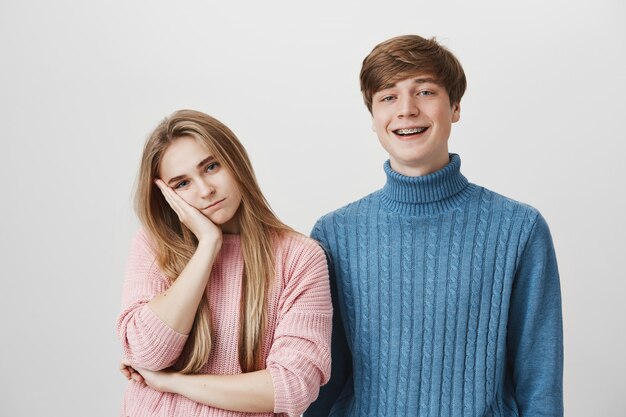 Dos personas de pie, chica se siente aburrida mientras chico sonriendo alegre