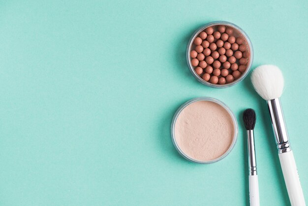 Dos pinceles de maquillaje con perlas compactas y bronceadas sobre fondo verde