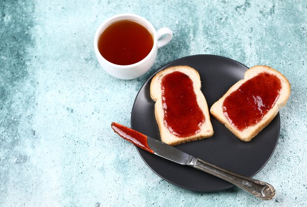 Dos rebanadas de pan con mermelada de fresa en un plato negro con una taza de té.