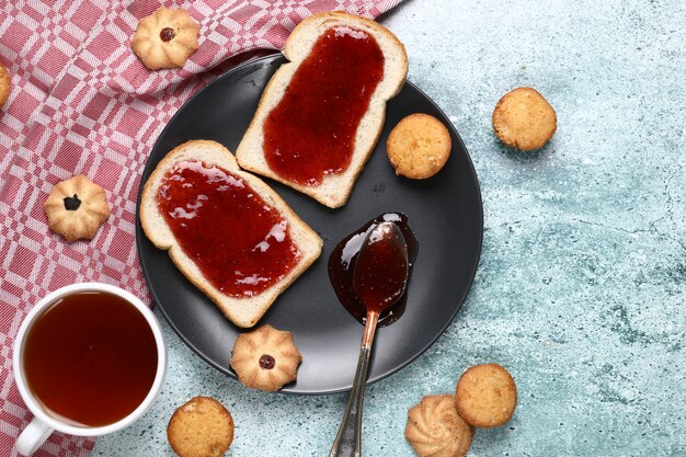 Dos rebanadas de pan tostado con mermelada roja en un plato negro con galletas alrededor