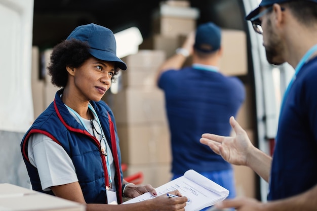 Dos repartidores se comunican y revisan el papeleo mientras sus colegas cargan paquetes en una camioneta. El foco está en la mujer afroamericana.