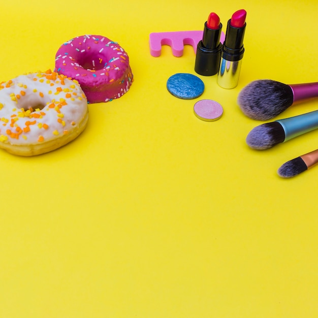 Foto gratuita dos rosquillas con lápiz labial; sombra de ojos y tres pinceles de maquillaje sobre fondo amarillo