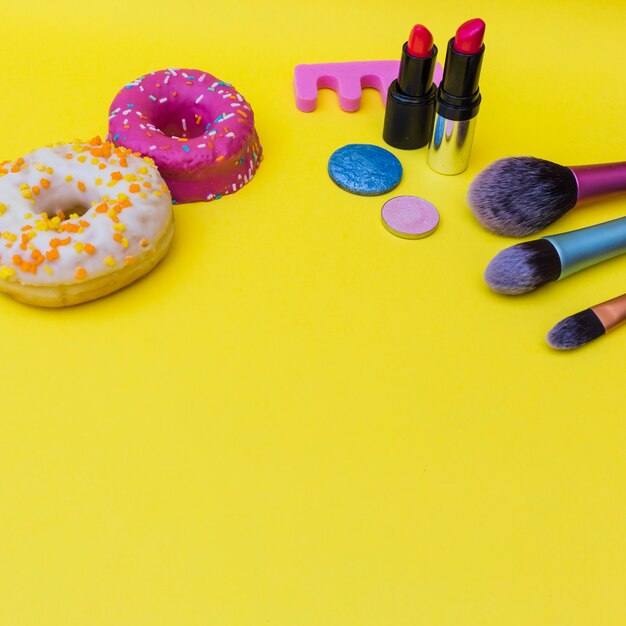 Dos rosquillas con lápiz labial; sombra de ojos y tres pinceles de maquillaje sobre fondo amarillo