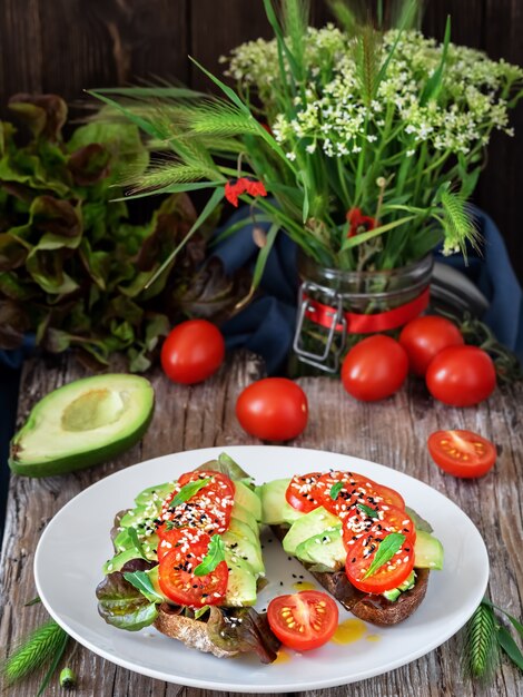 Dos sándwiches con pan recién horneado, aceite de oliva, lechuga, aguacate, semillas de sésamo y tomates cherry en una placa blanca.