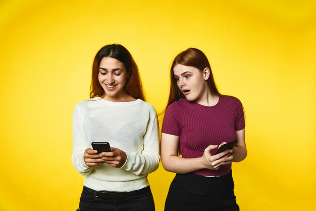 Dos sonrientes chicas caucásicas con teléfonos inteligentes modernos están mirando en la pantalla del teléfono