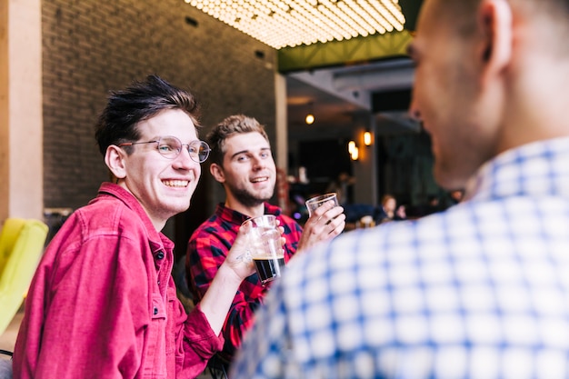 Dos sonrientes jóvenes sosteniendo vasos de cerveza mirando a su amigo