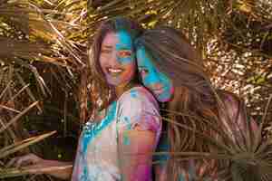 Foto gratuita dos sonrientes mujeres jóvenes con color holi en la cara mirando a la cámara