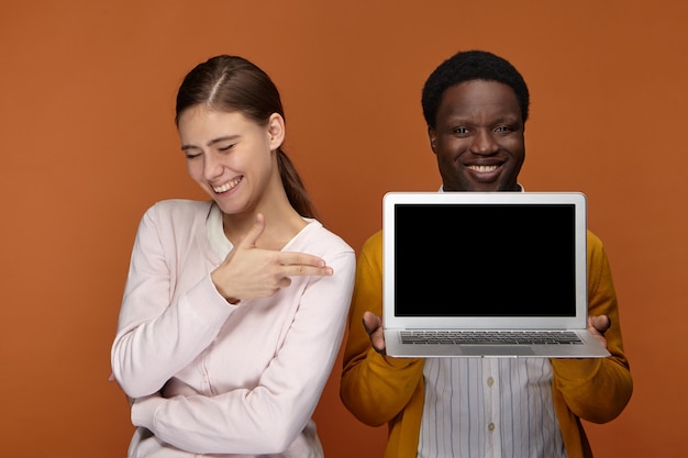 Dos talentosos jóvenes profesionales que disfrutan trabajando juntos en equipo: alegre hombre negro confiado que sostiene una computadora portátil genérica mientras una mujer blanca linda positiva apuntando a la pantalla en blanco, mostrando la presentación