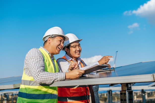 Foto gratuita dos técnicos e ingenieros asiáticos usan una computadora portátil mientras verifican la eficiencia del sol para examinar la construcción de paneles solares y hablar juntos