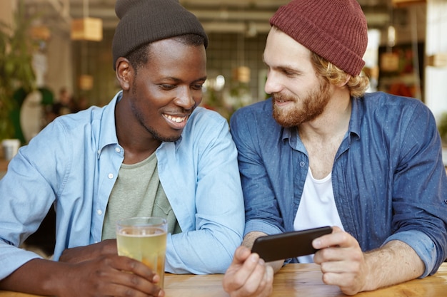 Dos tipos de diferentes razas bebiendo cerveza en el pub. Chico blanco de aspecto moderno con barba espesa que tiene una conversación agradable con su amigo