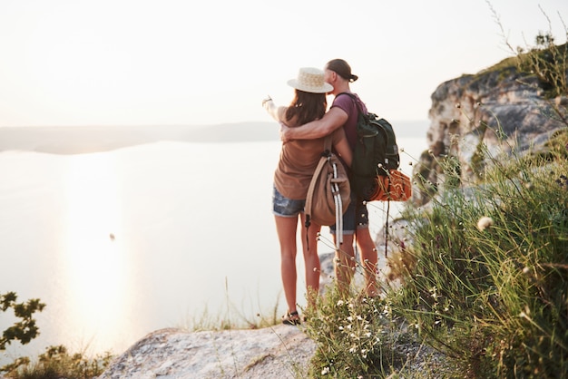 Dos turistas hombres y mujeres con mochilas se colocan en la cima de la peña y disfrutan del amanecer.