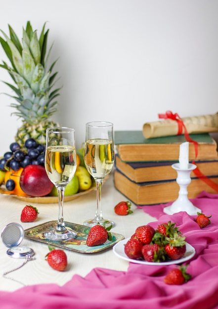 Dos vasos de champaña en la mesa de madera blanca con libros antiguos y reloj, diferentes frutas tropicales y fresas