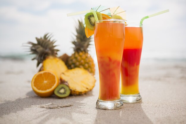 Dos vasos de cóctel de bebidas y frutas tropicales mantienen en la arena