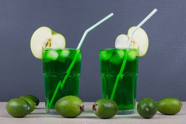 Dos vasos de jugo verde con pajitas en la pared azul.