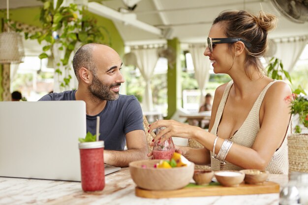 Dos viejos amigos que tienen una conversación animada, sentados a la mesa con un portátil y bebidas, contentos de verse después de mucho tiempo.