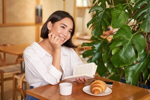 Foto gratuita dreamy young smiling asian woman reading book sitting in cafe eating croissant and drinking coffee i