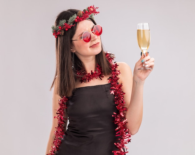 Dudosa joven bastante caucásica vestida con corona de Navidad y guirnalda de oropel alrededor del cuello con gafas sosteniendo y mirando una copa de champán aislado sobre fondo blanco.