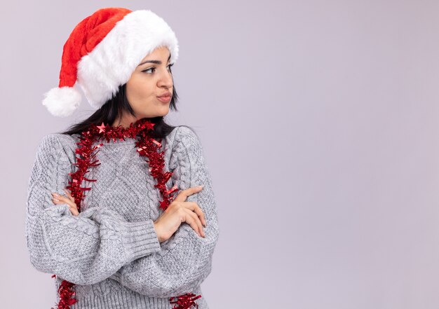 Dudosa joven caucásica vistiendo gorro de navidad y guirnalda de oropel alrededor del cuello de pie con postura cerrada mirando al lado aislado en la pared blanca con espacio de copia