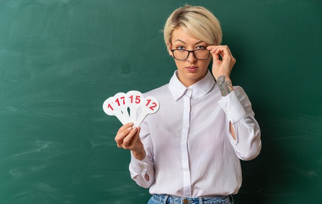 Dudosa joven profesora rubia con gafas en el aula de pie delante de la pizarra mostrando el número de ventiladores agarrando gafas mirando al frente con espacio de copia