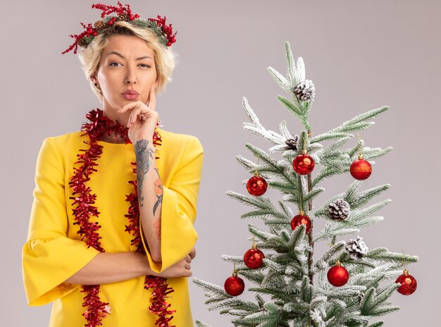 Dudosa joven rubia con corona de Navidad y guirnalda de oropel alrededor del cuello de pie cerca del árbol de Navidad decorado mirando manteniendo la mano en la barbilla aislada en la pared blanca