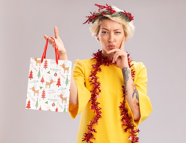 Dudosa joven rubia con corona de Navidad y guirnalda de oropel alrededor del cuello sosteniendo una bolsa de regalo de Navidad mirando manteniendo la mano en la barbilla aislada en la pared blanca