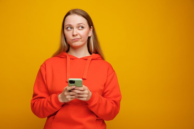 Dudosa joven rubia sosteniendo teléfono móvil mirando hacia arriba con los labios fruncidos