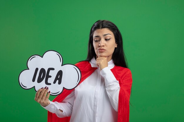 Dudosa joven supermujer sosteniendo la burbuja de la idea y mirándola con sospecha mirando directamente aislado en la pared verde