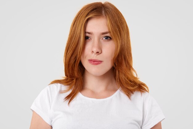 La dudosa mujer bonita frunce los labios y mira con vacilación, tiene el cabello rojizo, se viste con una camiseta informal, trata de elegir