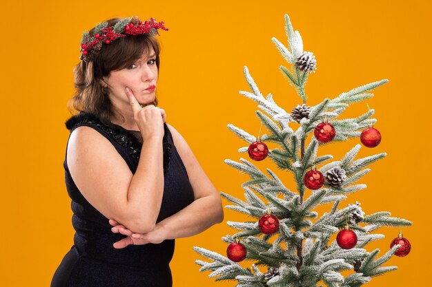 Dudosa mujer de mediana edad con corona de cabeza de navidad y guirnalda de oropel alrededor del cuello de pie cerca del árbol de navidad decorado en vista de perfil tocando la cara