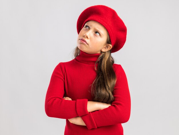 Dudosa niña rubia vistiendo boina roja manteniendo los brazos cruzados mirando hacia arriba aislado en la pared blanca con espacio de copia