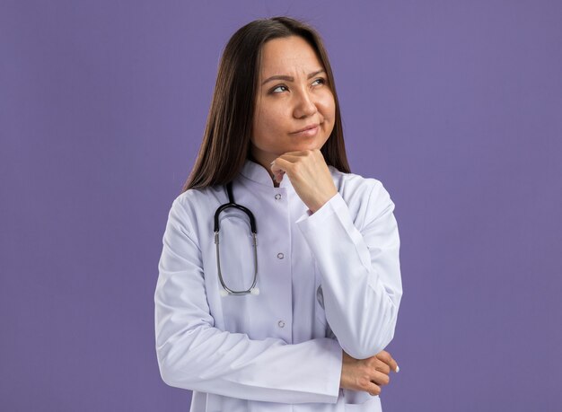 Dudoso joven doctora asiática vistiendo bata médica y estetoscopio manteniendo la mano en la barbilla mirando hacia arriba aislado en la pared púrpura con espacio de copia