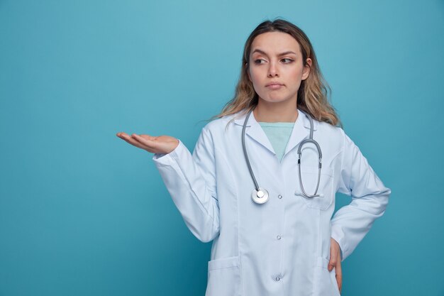 Dudoso joven doctora vistiendo bata médica y un estetoscopio alrededor del cuello manteniendo la mano en la cintura mostrando la mano vacía mirando al lado