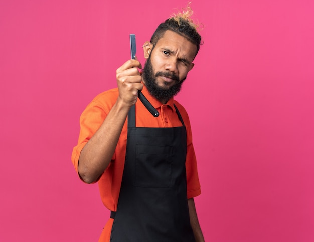 Foto gratuita dudoso joven peluquero masculino afroamericano vistiendo uniforme mostrando navaja recta