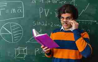 Foto gratuita dudoso joven profesor de geometría con gafas de pie delante de la pizarra en el aula mirando al frente sosteniendo un libro abierto agarrando gafas