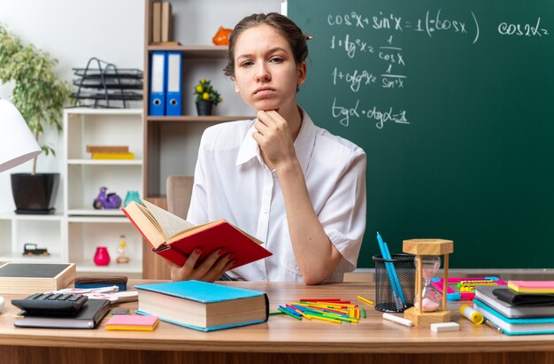 Dudoso joven profesora de matemáticas sentada en un escritorio con útiles escolares sosteniendo libros manteniendo la mano debajo de la barbilla mirando al frente en el aula