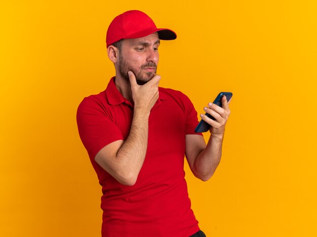 Dudoso joven repartidor caucásico en uniforme rojo y gorra manteniendo la mano en la barbilla sosteniendo y mirando el teléfono móvil aislado en la pared naranja