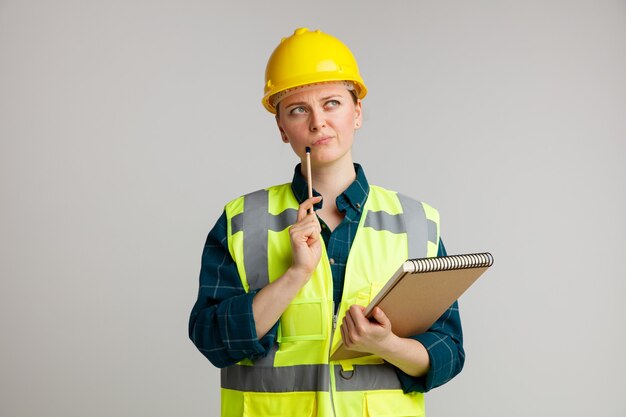 Dudoso joven trabajador de la construcción con casco de seguridad y chaleco de seguridad sosteniendo el bloc de notas tocando la barbilla con un lápiz mirando hacia arriba