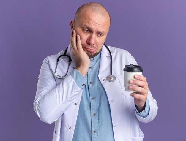 Dudoso médico varón de mediana edad vistiendo bata médica y estetoscopio sosteniendo y mirando una taza de café de plástico manteniendo la mano en la cara