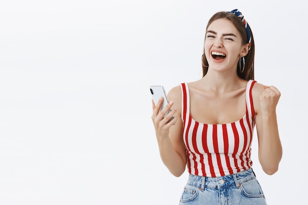 Dueña de la tienda en línea femenina celebrando la gran oferta en línea sosteniendo el teléfono inteligente apretando encaja en el gesto de éxito gritando sí, feliz y alegre celebrando la victoria