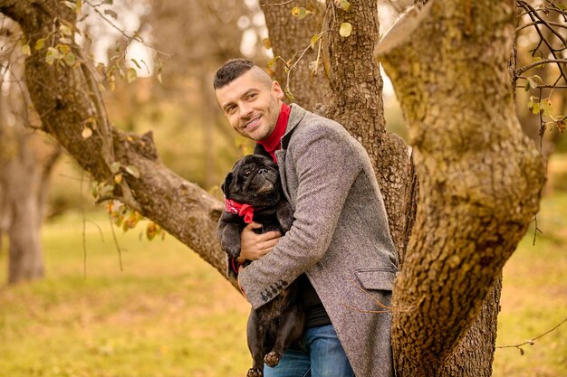 Dueño cariñoso. Un hombre de pie cerca del árbol con un perro en las manos.