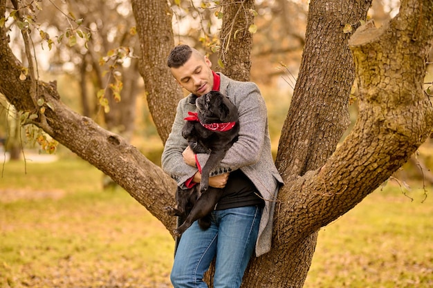 Dueño cariñoso. Un hombre de pie cerca del árbol con un perro en las manos.