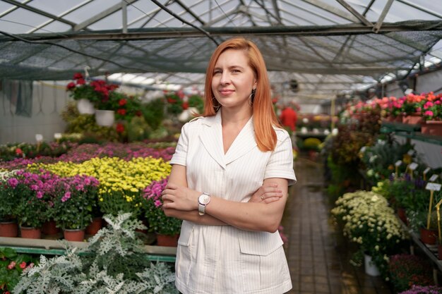 Dueño de invernadero posando con los brazos cruzados con muchas flores y un colega sosteniendo una maceta con crisantemos rosados bajo techo de vidrio