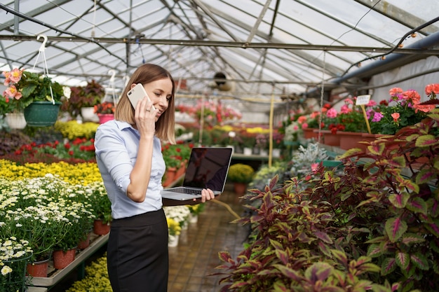 Dueño de invernadero sonriente posando con una computadora portátil en sus manos hablando por teléfono con muchas flores y techo de cristal.