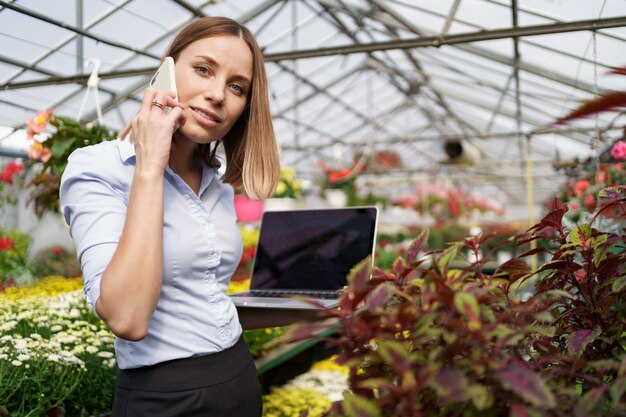 Dueño de invernadero sonriente posando con una computadora portátil en sus manos hablando por teléfono con muchas flores y techo de cristal.