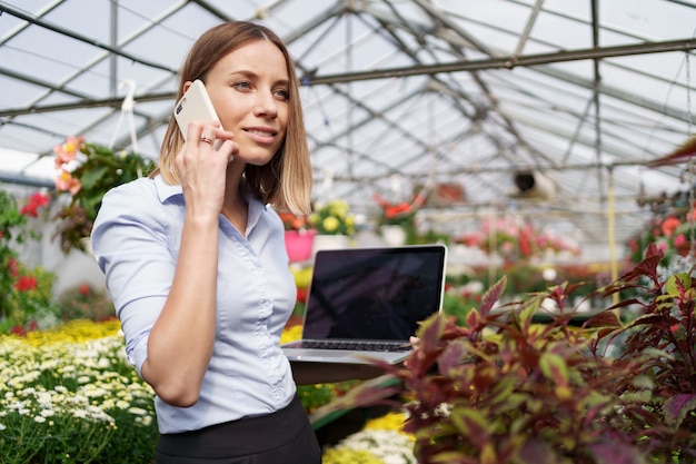 Dueño de invernadero sonriente posando con una computadora portátil en sus manos hablando por teléfono con muchas flores y techo de cristal.