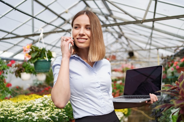 Dueño de invernadero sonriente posando con una computadora portátil en sus manos hablando por teléfono con muchas flores y techo de cristal.